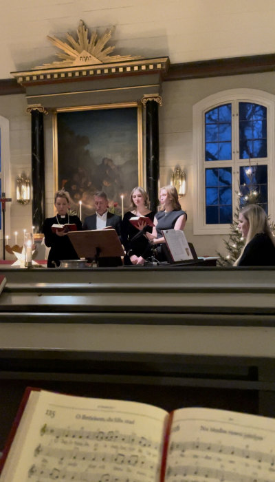 Kör sjunger i kyrka, notblad i förgrunden, adventsljusstakar, tavla på väggen, notställ, fönster med utsikt.