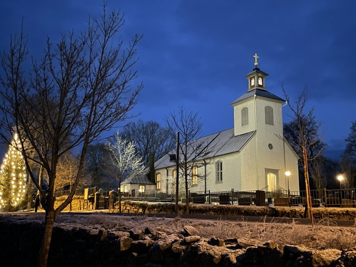 Vit kyrka om kvällen, julgran, belysning, barträd, stenmur, blå himmel, lugn atmosfär.