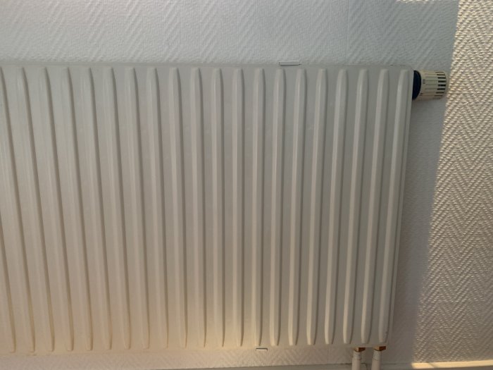 Vit värmeelement/radiator mot texturerad vägg, termostatventil synlig i höger hörn.