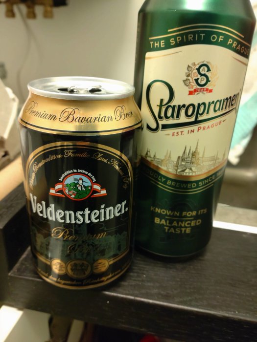 Två ölburkar, en Veldensteiner och en Staropramen, på en mörk yta, båda är stängda.