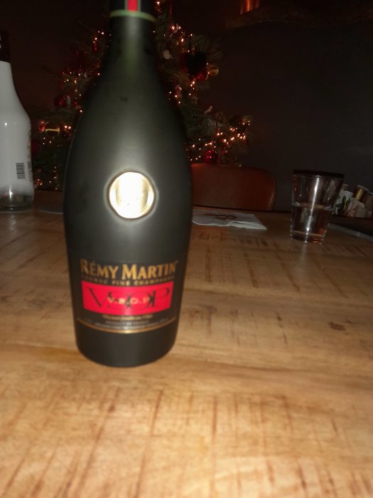 Flaska Rémy Martin VSOP på träbord. Julgran med ljus i bakgrunden. Oskarp, inomhus.