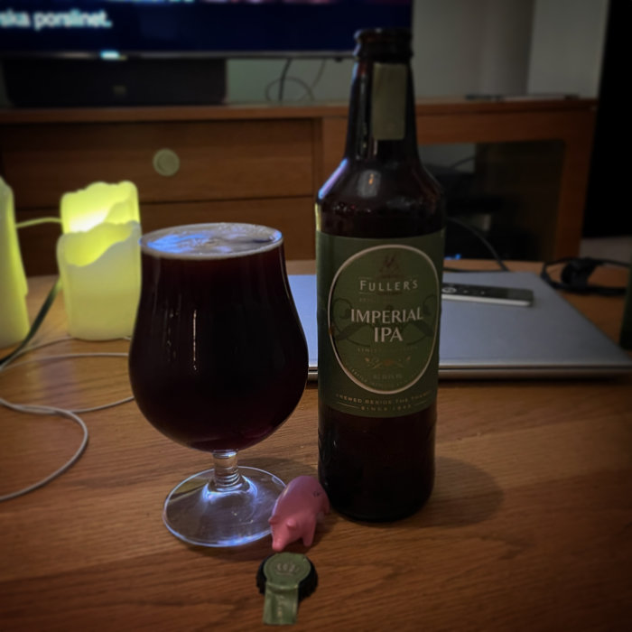 En ölflaska, glas fyllt med öl, bärbar dator och tv i bakgrunden, mysig atmosfär.