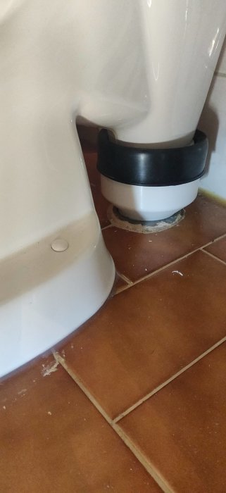 Toalettstol med avloppsanslutning ovanpå kaklat golv, tätningssvart ring syns, behov av städning.