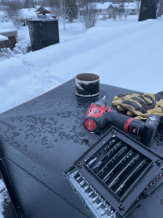Vinter, verktyg, borr, bitssats, handskar, mugg, utomhus på svart yta, snöigt bostadsområde i bakgrunden.