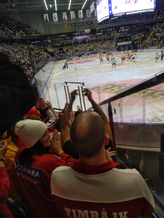 Hockeymatch pågår. Publik i klacksektion klädd i lagfärger. Spelare synliga på isen. Stor skärm ovanför.