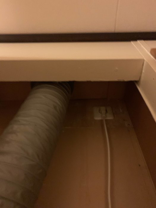 Flexibel ventilationskanal går genom tät öppning under vit möbel nära eluttag på trägolv.