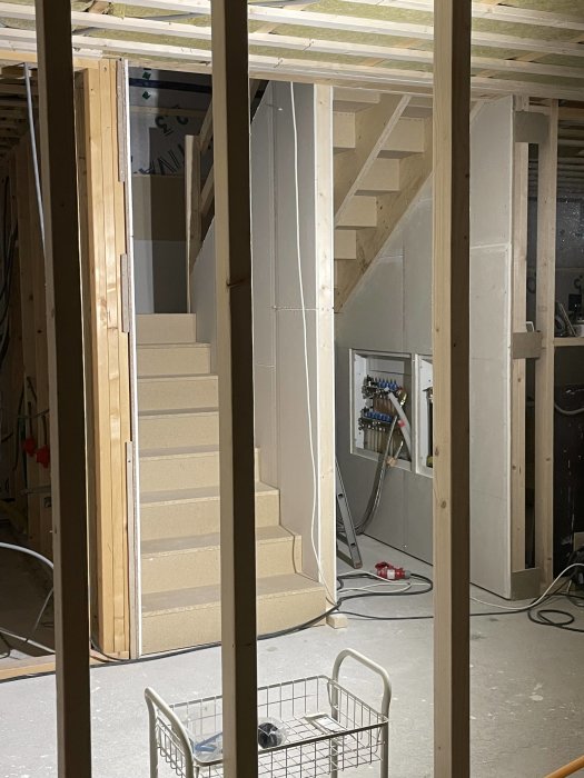 Inomhusbyggarbetsplats med trästommar, ofärdig trappa och elektriska installationer. En tom handlingsvagn i förgrunden.