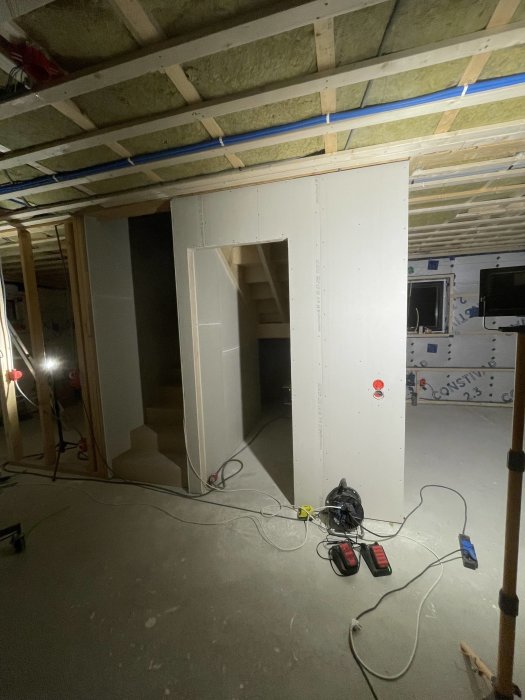 Byggarbetsplats inomhus med ofärdig gipsvägg, isolering i taket och verktyg på golvet.