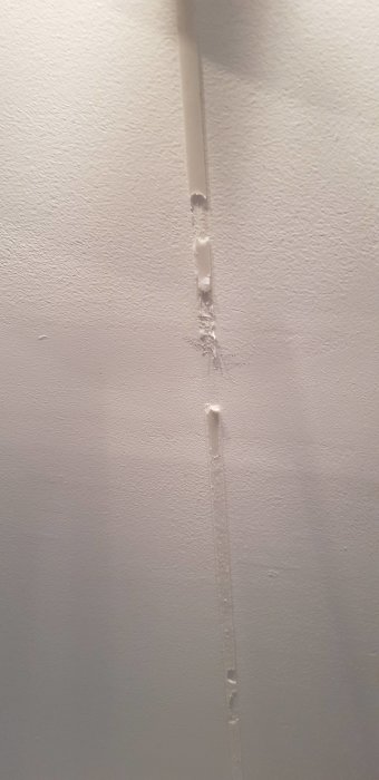 Vit vägg med skador, spikhål och slitage längs en vertikal linje.