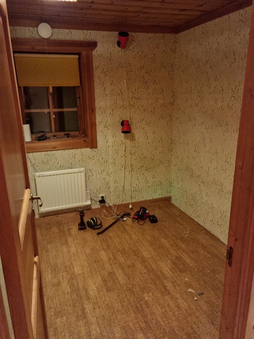 Ett tomt rum med träpaneler, mönstrad tapet, korkgolv, och verktyg på golvet.