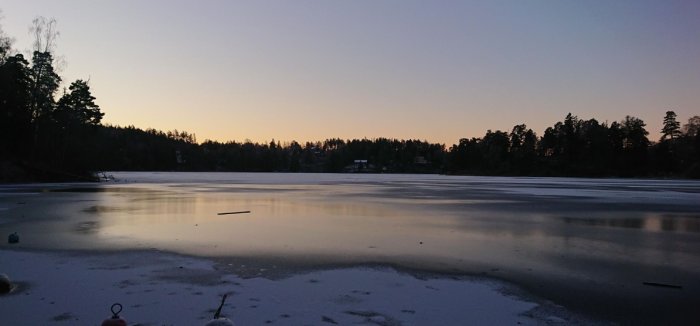 Skymning över ett frusset vatten med skog och hus i bakgrunden. Speglar himlen. Lugnt och kallt.