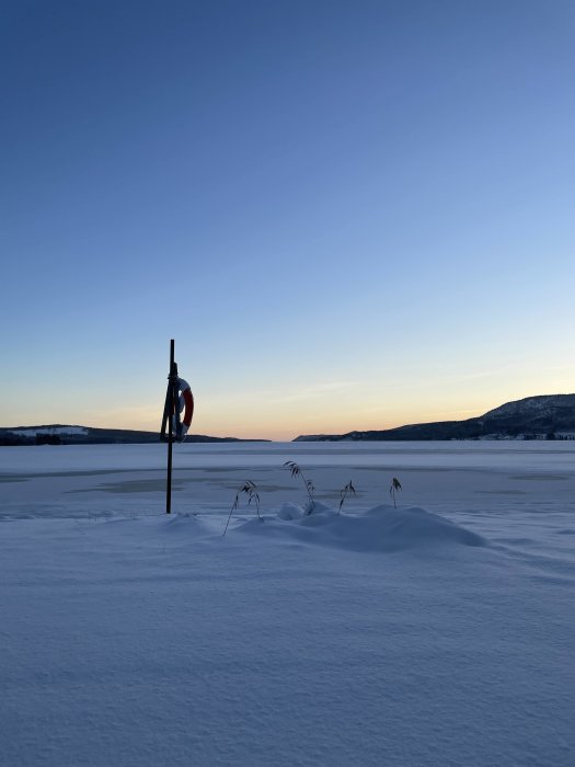 En frusen sjö vid solnedgång, med livbojställning och några snötäckta växter i förgrunden, klar himmel ovan.