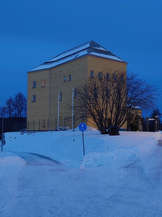 Gul byggnad i skymning, snö, kallt, gångvägsskylt, vinterlandskap.