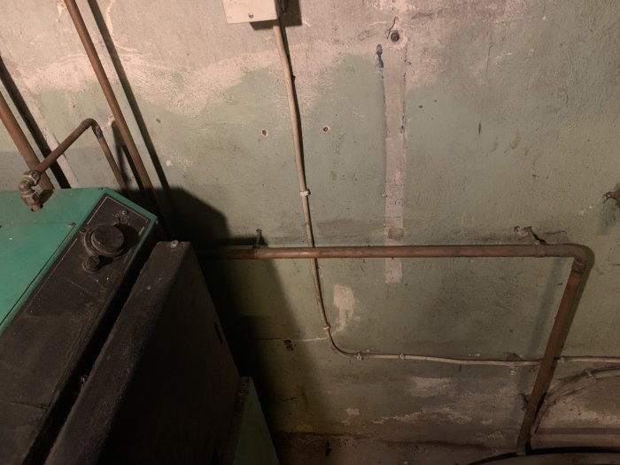 Äldre källarrum med rörledningar, slitna väggar och delar av en panna. Dustigt, omodernt och i behov av underhåll.