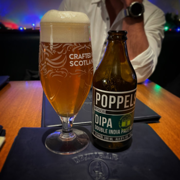 Dubbel IPA öl, glas, skummig topp, Poppels bryggeri, "Crafted Scotland", man i bakgrunden, mörk bordsyta.