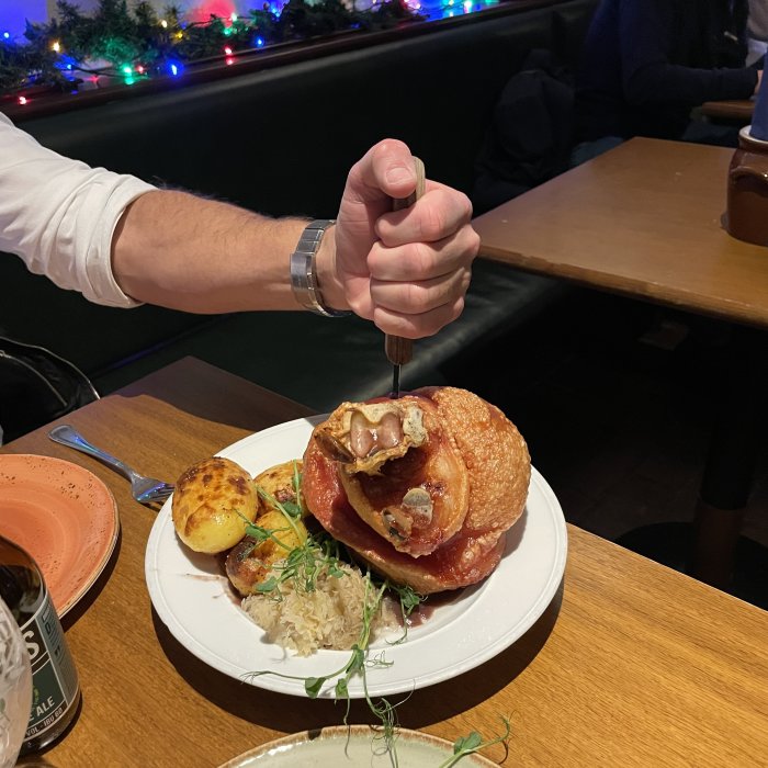 Person håller i kniv, skär i hamburgare med bacon, serverad med potatis och öl.