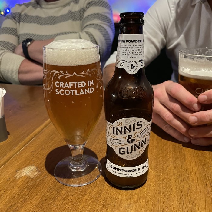 Ölglas märkt "Crafted in Scotland", Innis & Gunn flaska, personer i bakgrunden vid bardisk.