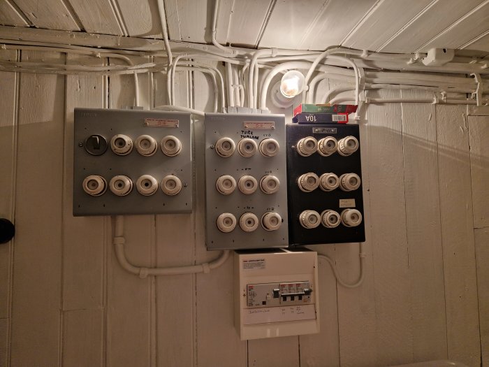 Elektriska säkringsskåp och strömbrytare på en vitmålad vägg, med synliga kablar och rör.