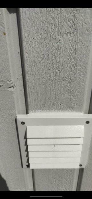 Vit ventilkåpa på texturerad vägg, solbelyst, skugga, skruvar synliga, modern, avlopps- eller ventilationselement.