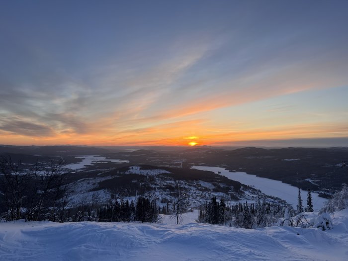 Vinterlandskap vid solnedgång, snöklädda backar, frusen sjö, skog, himmel med varma färger.