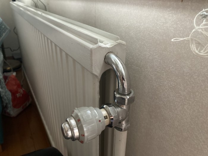 Radiator med termostatventil, vitmålad, mot en texturerad vägg, inomhusmiljö, diverse föremål i bakgrunden.