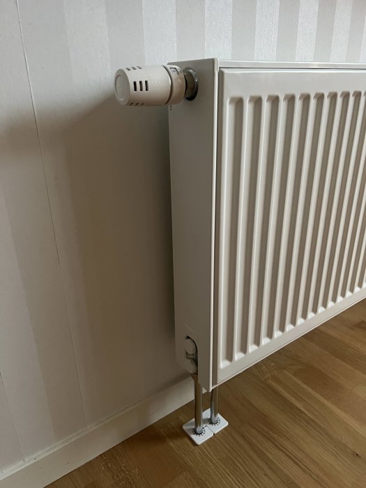 Väggmonterad radiator med termostatknopp, vit, inomhus, ventiler och rör synliga mot ljus tapet.
