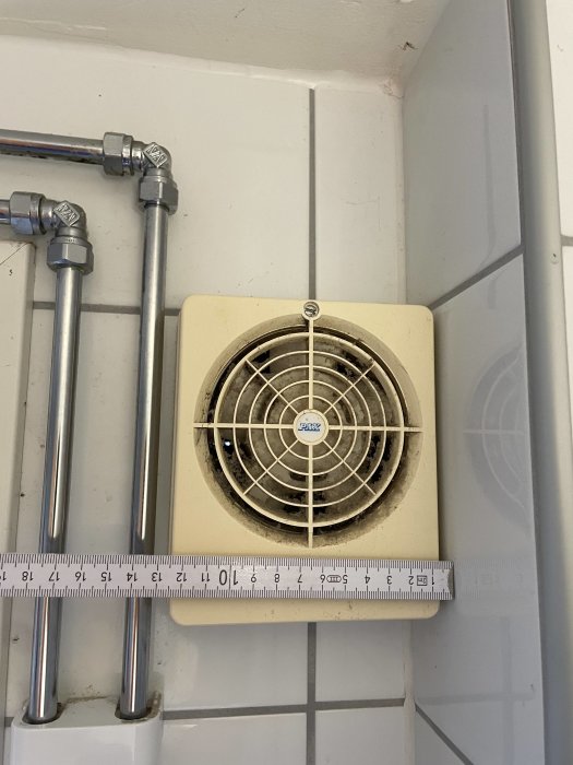 Ventilationsfläkt i vägg med skyddsgaller, rör och linjal för storleksreferens. Vit kakelbakgrund. Hygienutrymme möjligt.