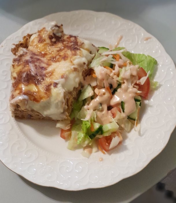 Bit lasagne med smält ost och sallad med rosa sås på vit tallrik.