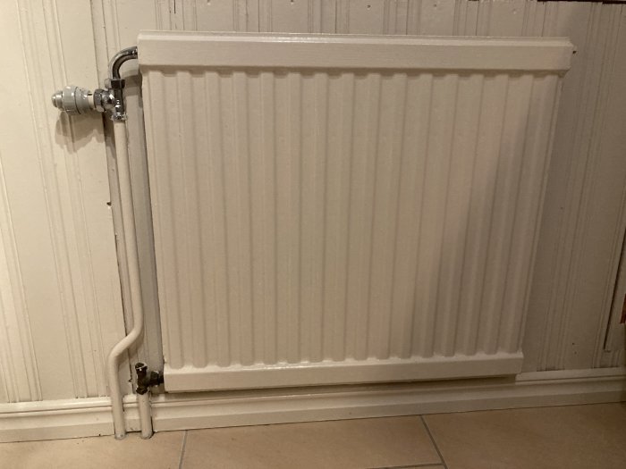 Vit elementvärmare vid vägg, termostatventil, rör, kaklat golv och panelvägg.