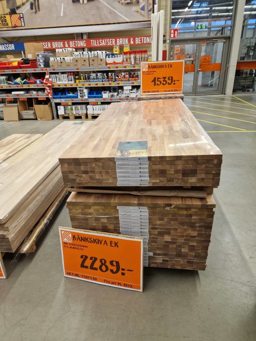 Staplade träbänkskivor med prisetiketter i byggmarknad. Belysning. Inga människor synliga.