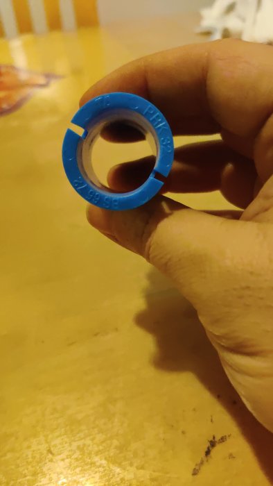 En hand håller en blå plastkapsyl med text; suddig bakgrund, inomhusmiljö.