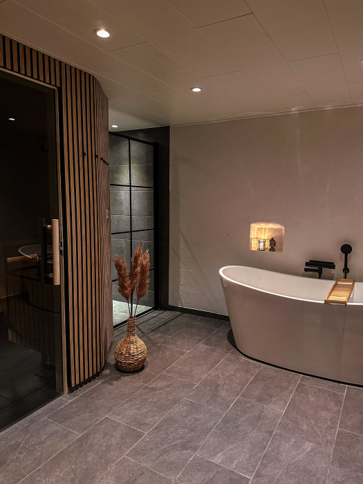 Modernt badrum, frittstående badkar, trädetaljer, gråa plattor, indirekt belysning, växtarrangemang, spegel och duschhörna.