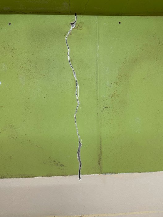 En stor spricka på en grönmålad betongvägg övergående till en beige yta nedtill. Slitage och smuts syns.