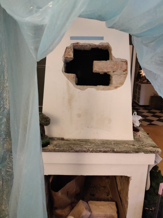 En öppen spis under renovering med synliga tegelstenar, täckt av plast, i ett rum med schackrutigt golv.