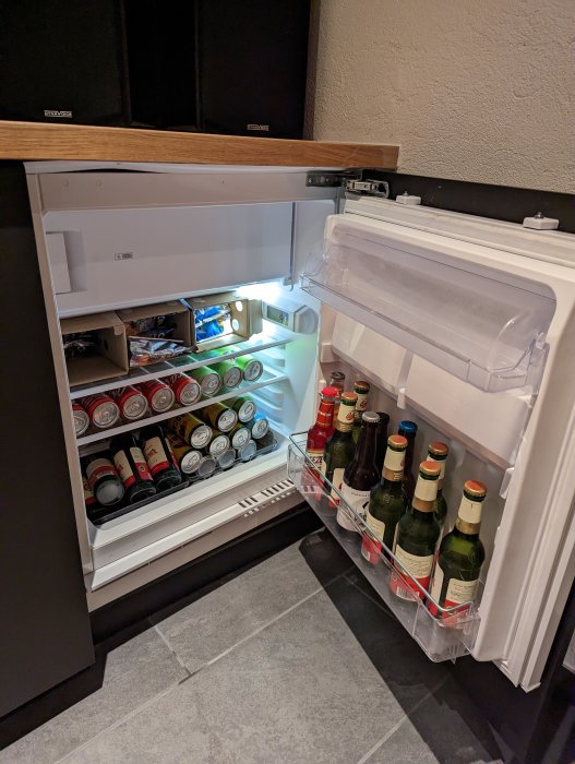 Ett öppet kylskåp fullt med burkar och flaskor, främst öl och läsk, inbyggt i köksskåp.