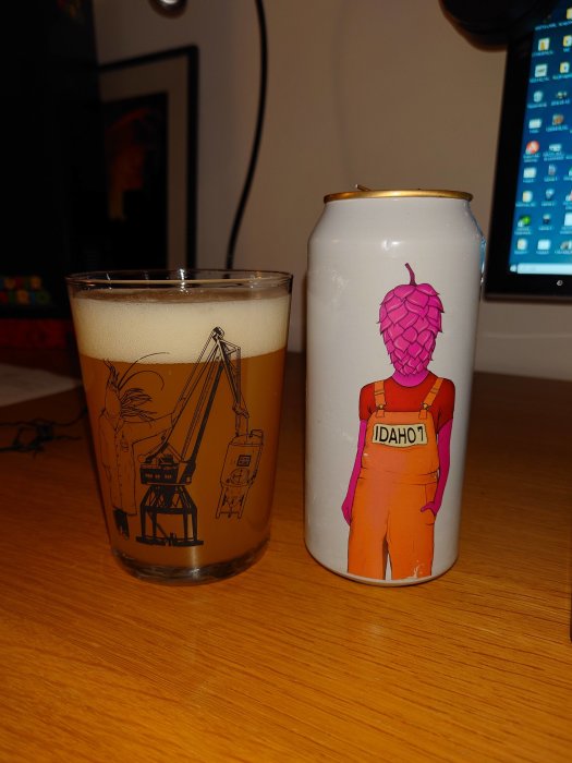 Ölglas med illustration, ölburk med humlekotte-design, bryggeriutrustning, dator i bakgrunden, träyta.