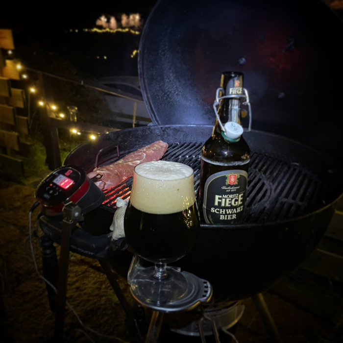 Kvällsgrillning med stek, ölflaska, skummande ölglas, och tänd grill omgiven av ljusslingor.