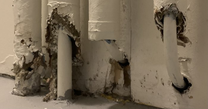 Gamla, skadade vattenrör i vägg, med flagnande färg och korrosionsskador. Behov av renovering synligt.