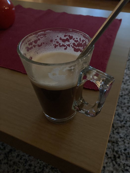Glasskopp med kaffe och skum, metallsked, träbord, röd duk, otydlig röd bakgrundsföremål.