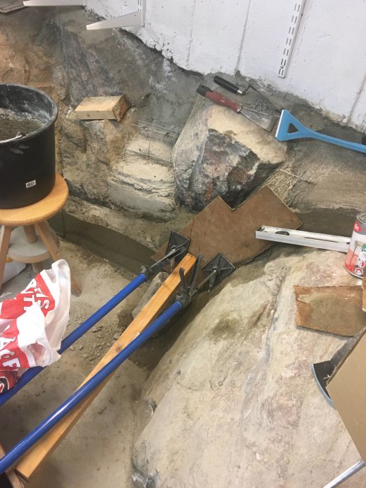Rörigt byggarbetsutrymme med verktyg, byggmaterial, klämmer och oslipad betongvägg.