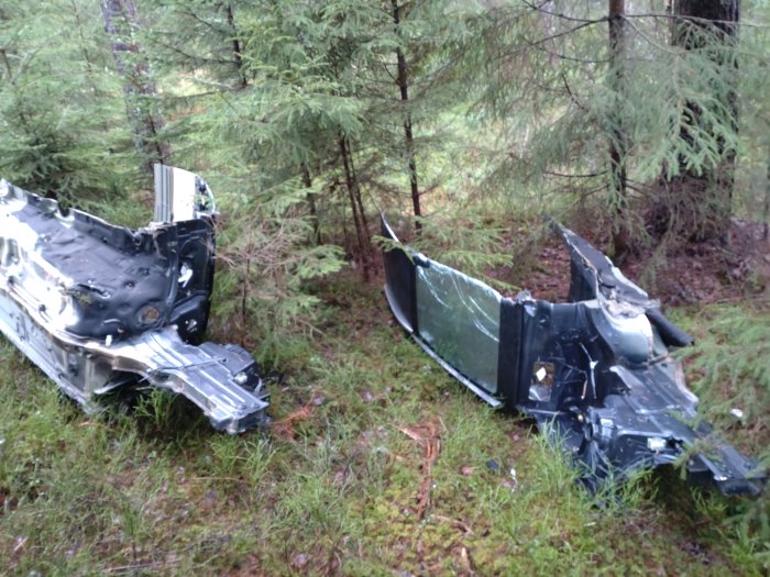 Kraftigt krockad bil delad i två, utspridd i skog. Skadad, övergiven, natur. Nedfallna grenar och mossa synlig.