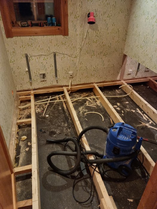 Ett pågående renoveringsrum med exponerat golvbjälklag, elinstallationer, tapet och en dammsugare.