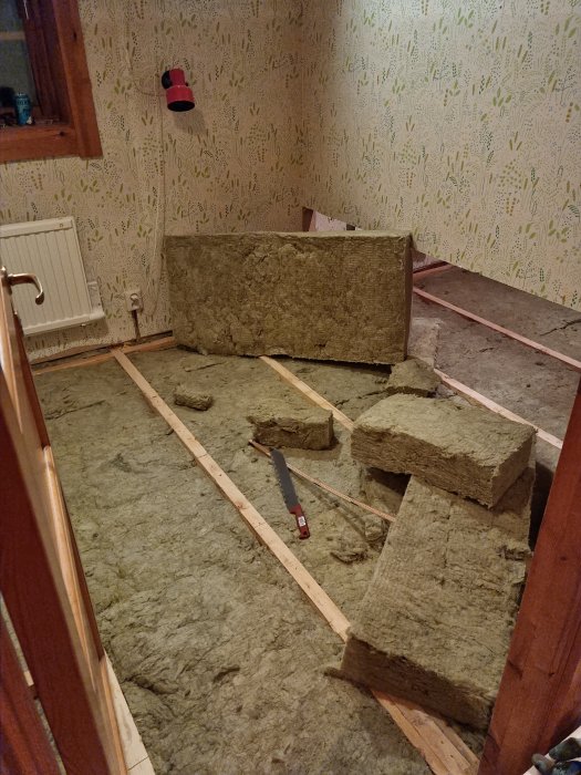 Renovering av rum, isoleringsmaterial på träbjälklag, vägg med tapet, handsåg och måttband synliga.