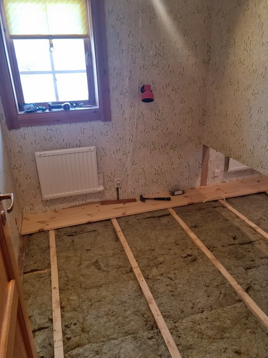 Renoveringsprojekt, tomrum, träreglar på isolerat golv, tapetserad vägg, fönster, radiator, arbetslampa.