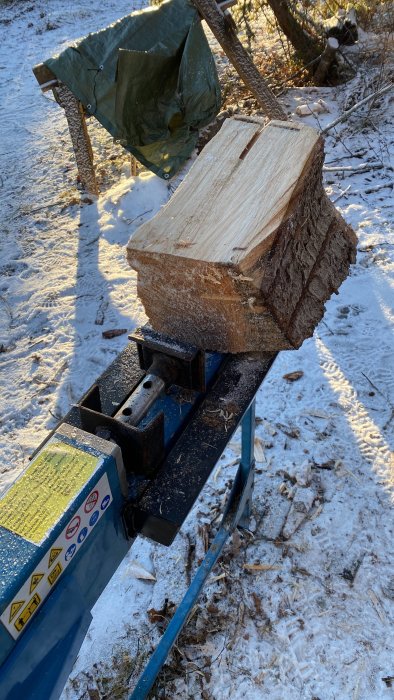 Stort träblock på vedklöv, utomhus, snö, vinterdag, skogsmiljö, klyvverktyg, blått, soligt, skuggor, kallt.