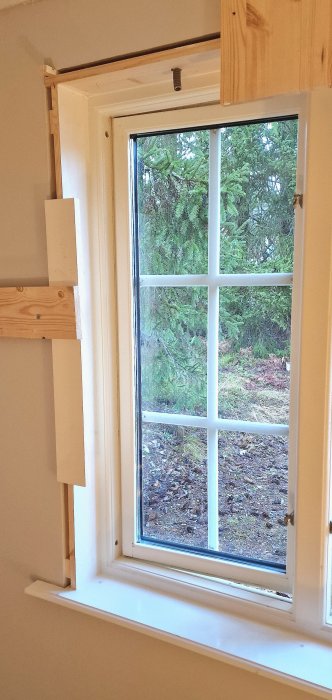 Ett vitmålat fönster, med träramar, som vetter mot gröna träd, under pågående konstruktionsarbete eller renovering.
