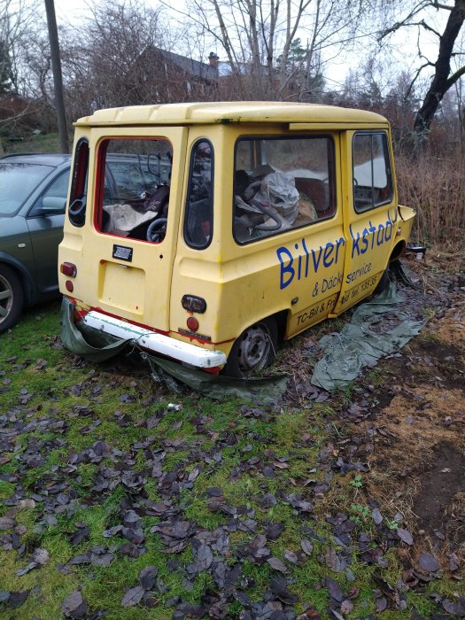 Gammal, övergiven bil med text, skräp inuti, skadad stötfångare, parkerad på gräsmark med löv.