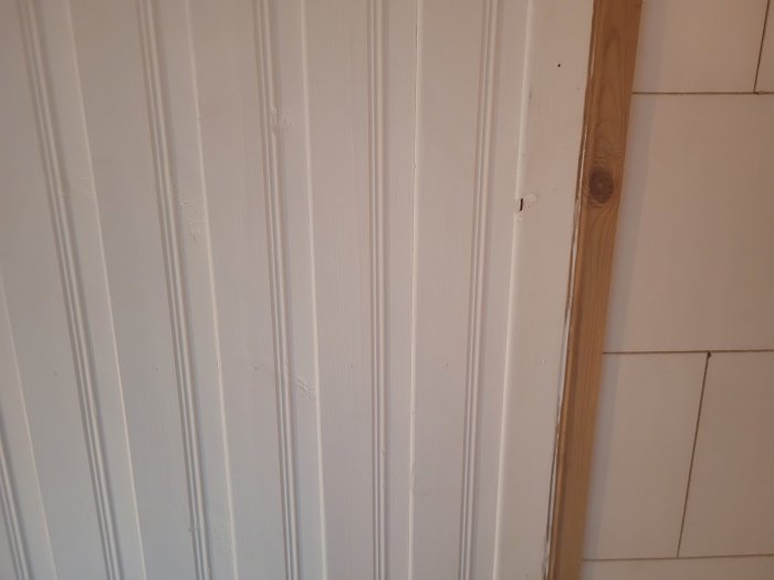 Vit panelvägg möter vit kaklad vägg, trälist i hörnet, texturkontrast, enkel heminteriör.