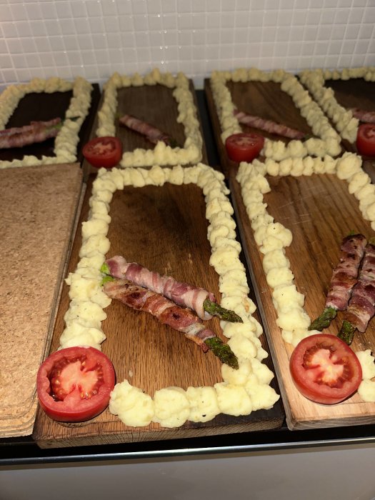 Träskärbrädor med potatismoskanter, baconlindade sparrisar, tomatdekoration, förberedelse för matpresentation.