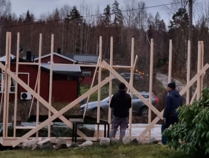 Två personer framför trästomme för byggnation, skymning, träd, hus och bil i bakgrunden.